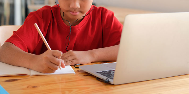 A boy with headphones in taking notes and looking at his laptop.