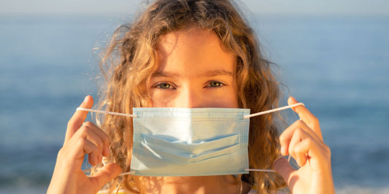 A girl holding a medical mask up to her face while on the beach.