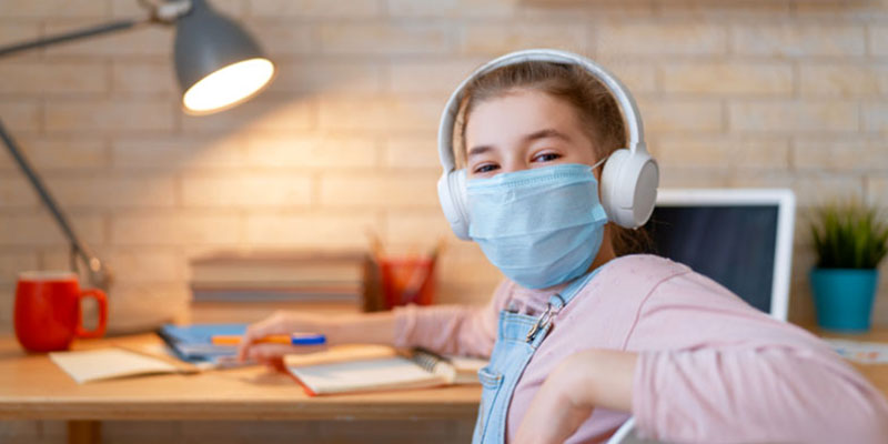 A girl wearing a facemask sitting at her home desk with the laptop open
