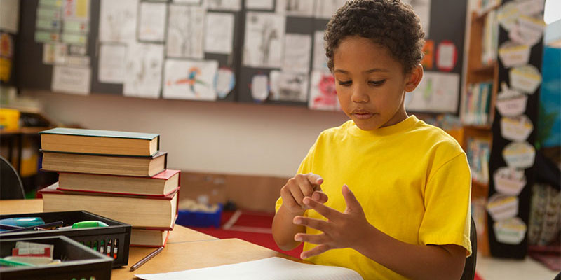 A young boy sits at a desk in a classroom counting on his fingers.