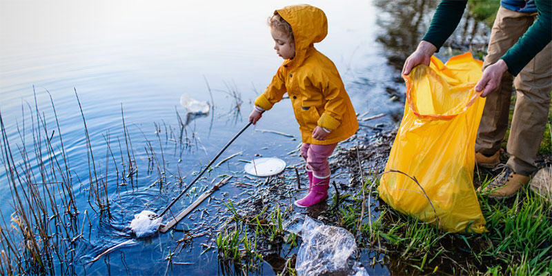 A young girl in a yellow raincoat uses a grabber to pick trash out of a pond while her parent holds open a trashbag.